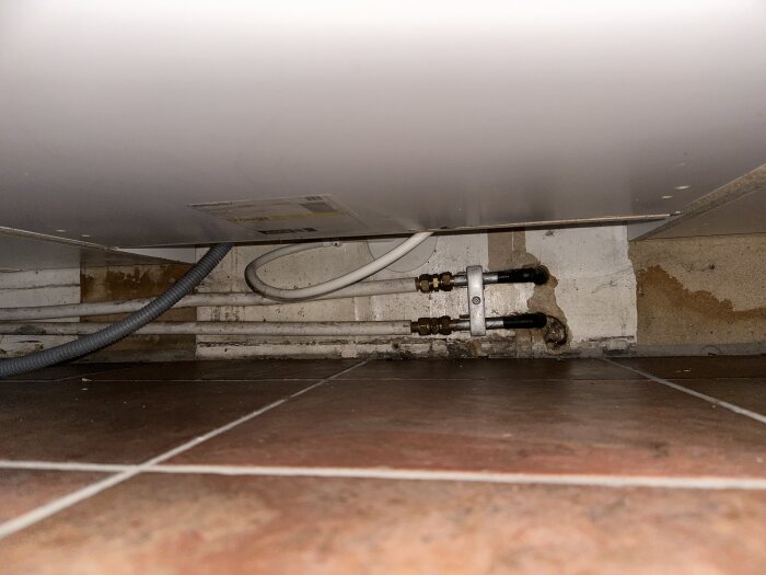 Vattenskada under köksspis med synliga rör och vattenfläckar på väggen. Problem med läckage från badrummet ovan som påverkar källartaket.