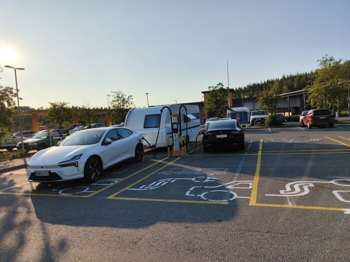 Två elbilar står parkerade och laddas vid en laddstation i Finland under en solnedgång, med omgivande skog och byggnader i bakgrunden.