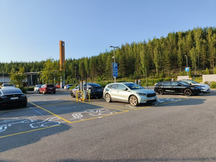 Bilar parkerade vid en laddstation i Finland, omgivna av träd och klarblå himmel.