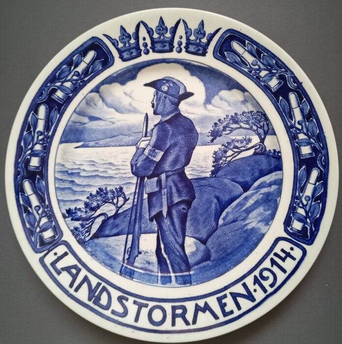 Tallrik med blåvit illustration av en soldat med gevär vid kustlandskap. Texten "Landstormen 1914" samt kungliga kronor syns i kantdekoren.