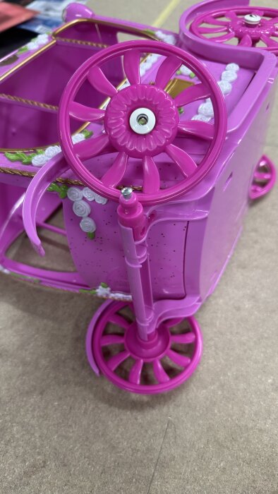 Rosa leksaksvagn med trasiga hjul. Blommor dekorerar kanterna på vagnen. Ett hjul har en synlig skruv i mitten.