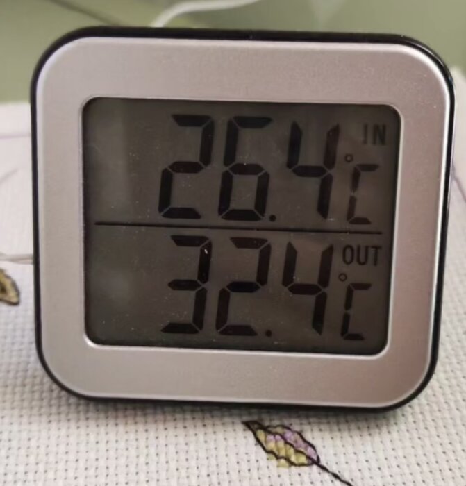 Digital termometer visar 26,4°C inomhus och 32,4°C utomhus, placerad på mönstrat tygunderlag.
