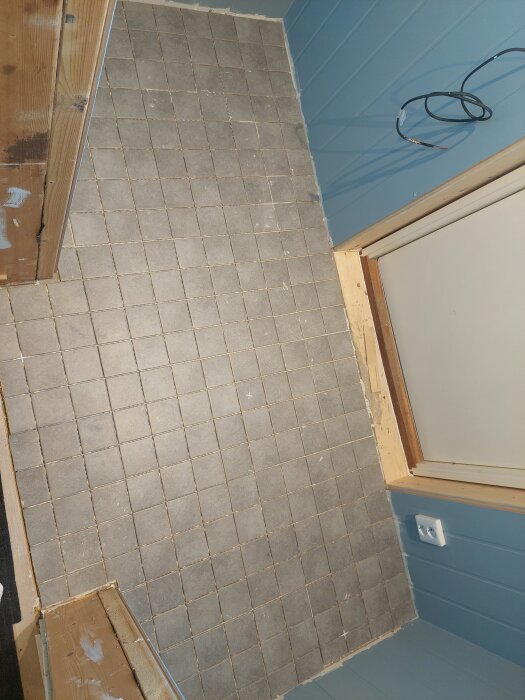 Klinkergolv i gråa kvadrater, del av oavslutat rum med blå väggar och trädetaljer. Sladd hänger från väggen nära en dörr.