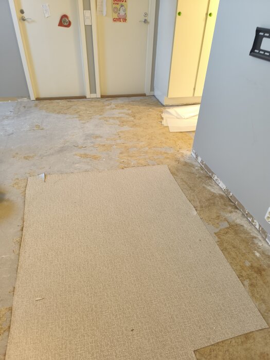 En beige plastmatta ligger ovanpå ett slitet golv i ett rum med vita dörrar och ljusblå väggar, diverse utrustning syns i hörnet.