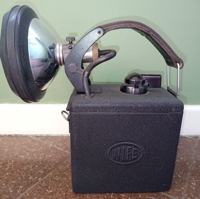 Handlampa från Jungnerbolaget med NiFe-batteri i metallhölje, placeras på klinkergolv framför grön vägg.