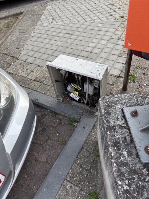 Skadat plastskåp utan lucka på en parkeringsplats i Belgien, med synliga kablage, nära en parkerad bil.