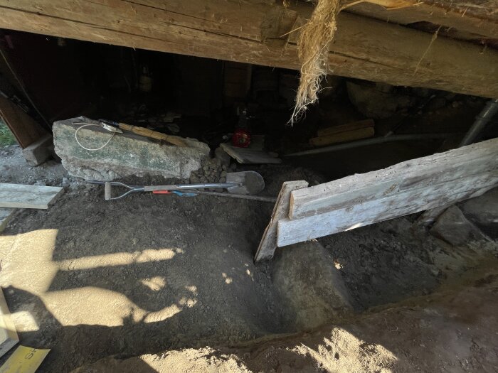 Jordgolv i gammal matkällare med stora stenar, ljus insläppt från ovan, trähylla för verktyg ovanpå. Grävverktyg och skrotade brädor synliga.