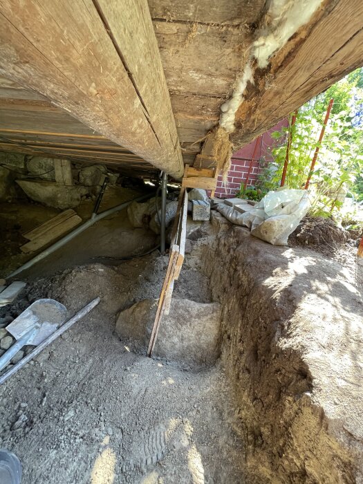 Under huset syns en stor stenbumling i jord, med träkonstruktion ovan och rör på sidan. Tomma plastpåsar och tegelvägg syns i bakgrunden.