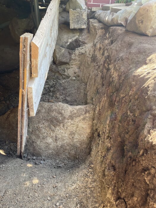 Utgrävd mark under torp med träskiva som skyddar avloppsrör, stora stenar synliga intill grävningsområdet.