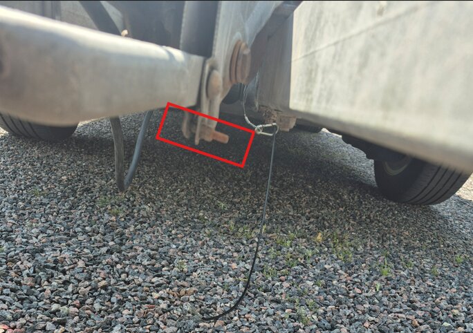 Släpvagnens undersida med bult och vajer, där vajern verkar ha lossnat. Markbeläggning är synlig under släpvagnen.