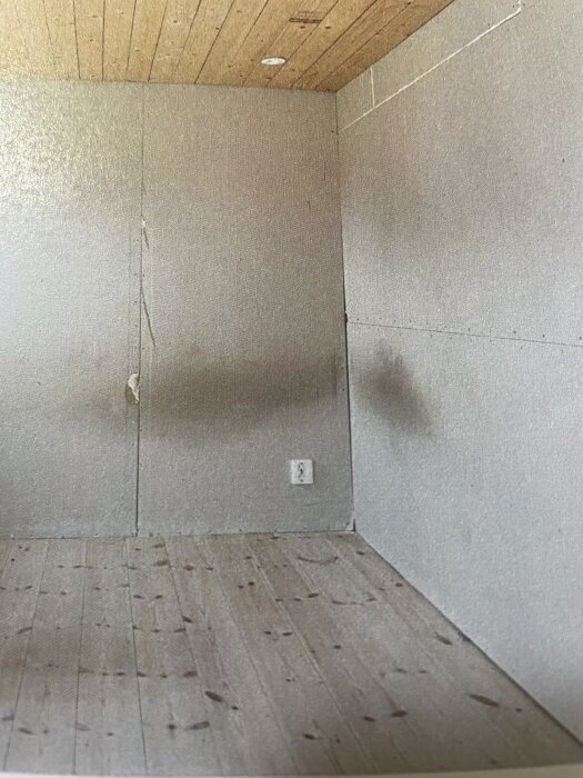 En ofärdig vägg i ett rum med synlig fläck på ytan och trägolv; taket är i trä. En kontaktuttag syns på väggen.