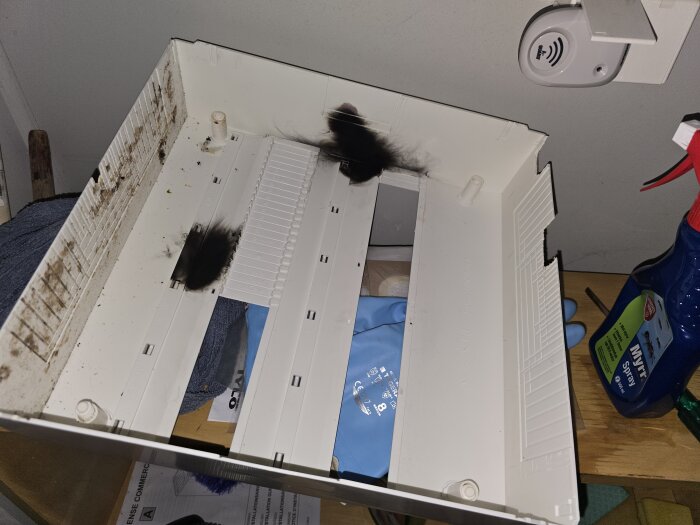 Sotiga märken inuti en plasthölje av en elcentral i en bastu, omgiven av rengöringsprodukter och en blå handske.