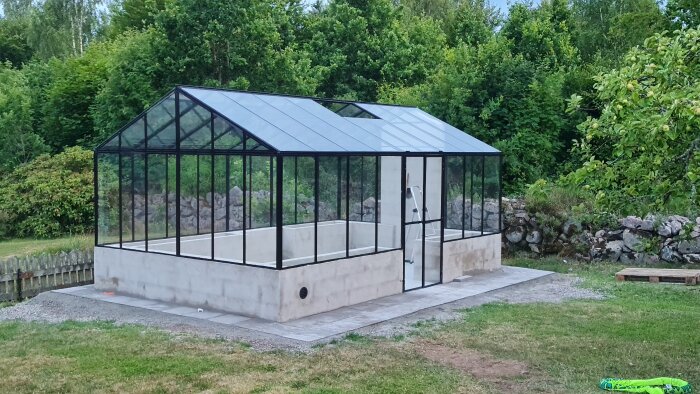 Nybyggt växthus med putsad mur där skarvar från lecablock syns igenom. Stege synlig på insidan, omgiven av gräsmatta och träd.