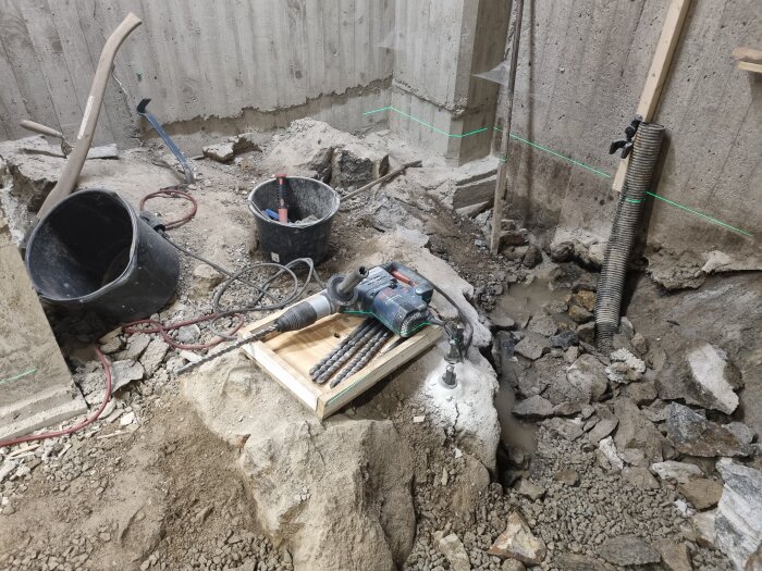 Bilden visar en byggarbetsplats med borrhammare, borrar, hinkar och stenar i ett hörn av en källare under renovering.