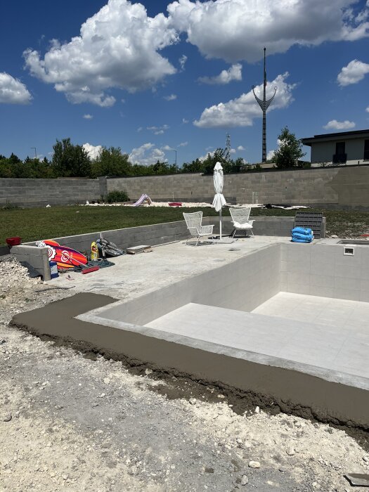 Poolområde under markarbete med grundläggande ytor för plattsättning, omgärdat av stolar och ett parasoll på en solig dag.