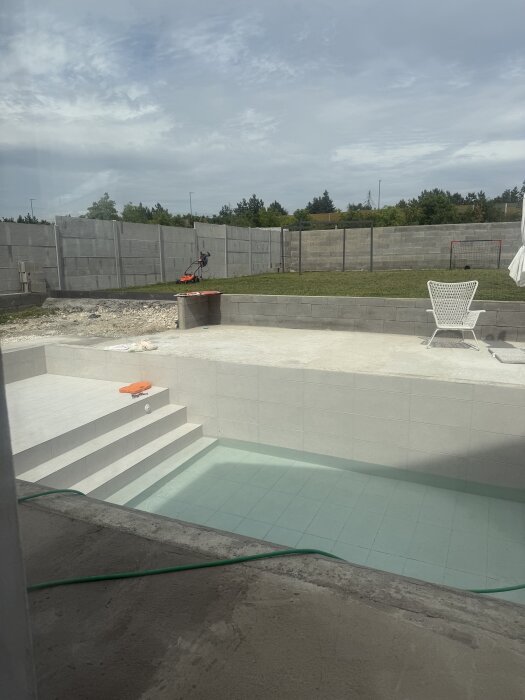 En halvfylld pool med vatten och en grön slang liggande vid poolens kant i en trädgård med betongväggar och en gräsmatta.