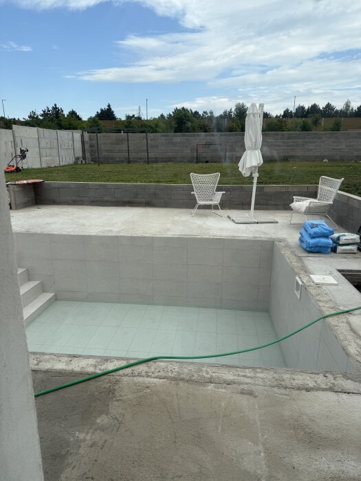 Fylld pool med vatten till en fjärdedel, omgiven av stenbelagd terrass, gräsmatta och utemöbler. Trädgårdslang fyller på vatten.