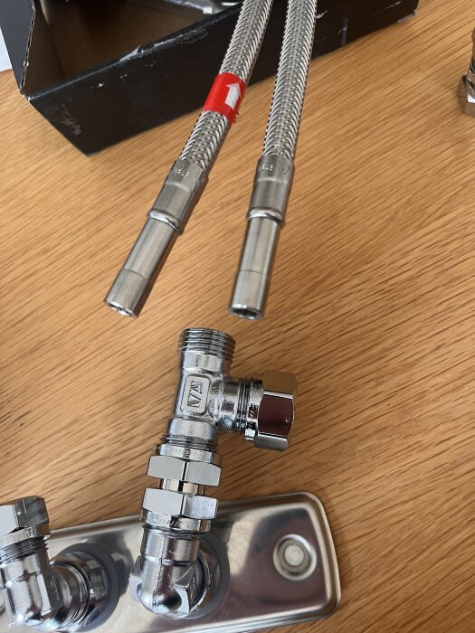 Slang och T-koppling i metall ligger på ett bord, med två flexibla rostfria slangar och en T-koppling med klämringskopplingar.