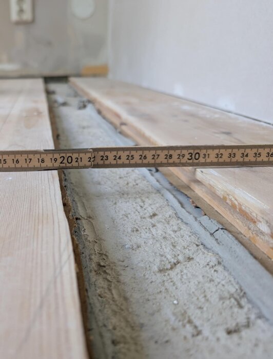 Öppet spår i golv med linjal, avslöjar betongunderlag där plankor saknas. Planeras att fyllas med golvspackel och matchande planka.