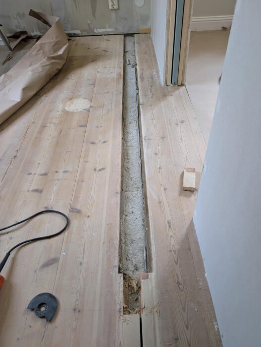 Renoveringsprojekt: Ett golv med en borttagen planka, synlig avjämningsmassa och verktyg på sidan. Rummet har furugolv och en buktande vägg.