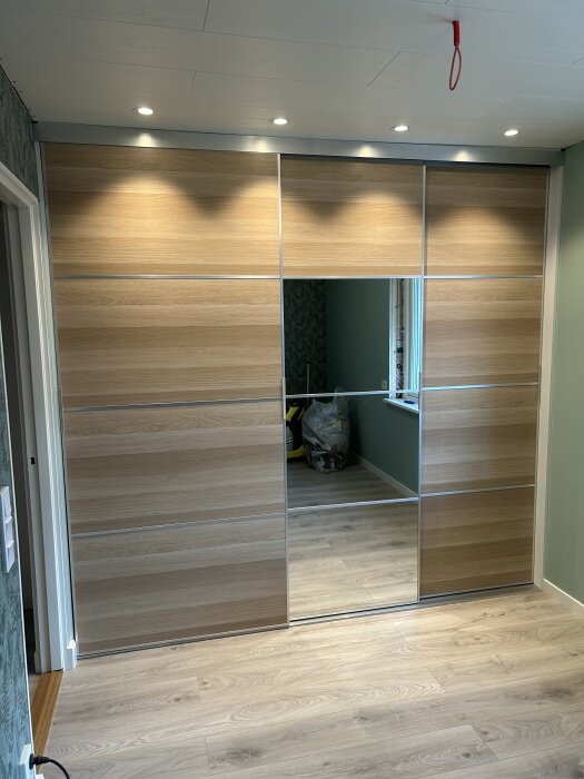 Inbyggd garderob med skjutdörrar i trä och spegel, takspottar ovanför, i ett rum med träparkett och grönmålade väggar.