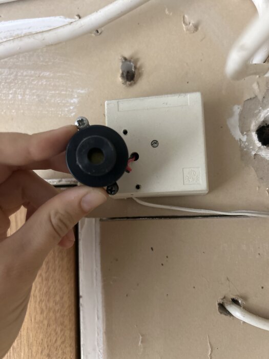 Hand som håller en svart, rund komponent framför en vit dosa på väggen med synliga kablar och borrade hål.