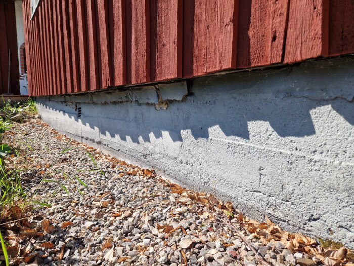 Röd stuga från 1957 med torpargrund där muren sjunker under huset. Grunden är skadad och omgiven av grus och löv.
