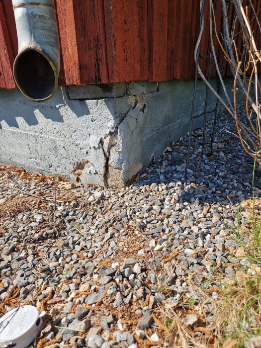 Sprucken husgrund med synlig skada på ett äldre torp, omgiven av rött trä och grusig mark. En hängränna ses till vänster.