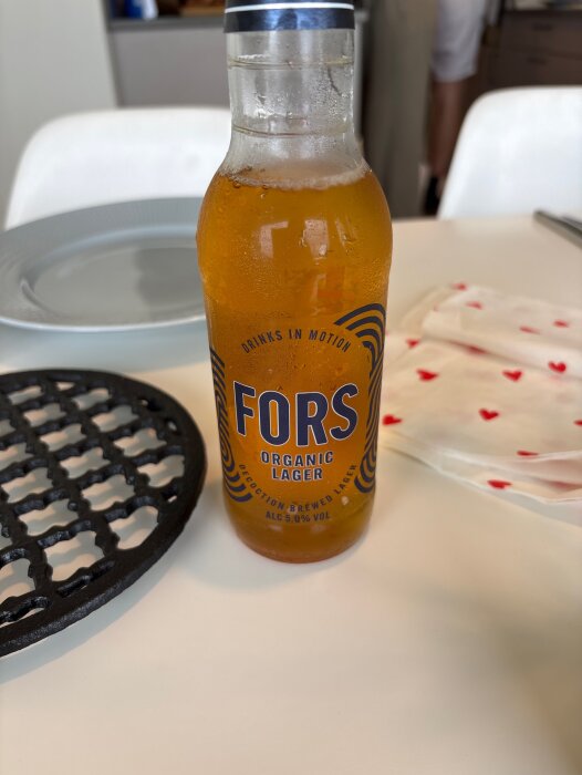 En flaska Fors Organic Lager står på ett bord bredvid ett svart grillgaller och en vit servett med röda hjärtan.