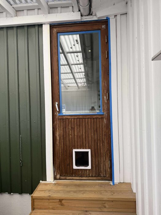 Ytterdörr med sliten träyta och hundlucka redo för ommålning, omgiven av vit och grön panel, ovanpå trätrappa.