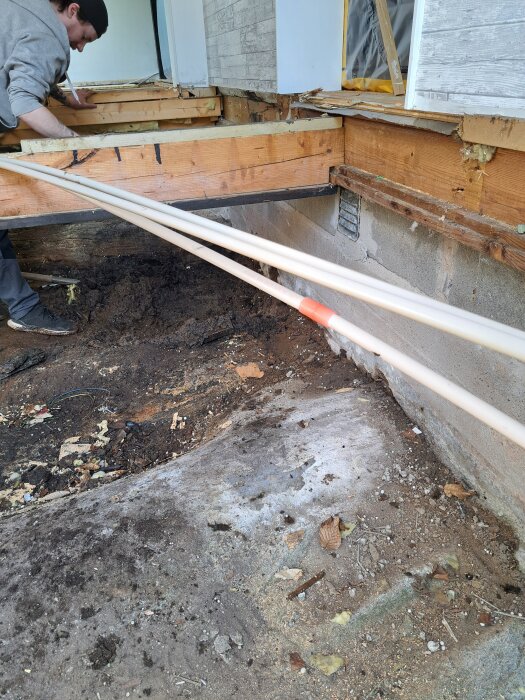 Skadad träkonstruktion med ruttna brädor, öppet förfall under ett hus, arbetare i keps inspekterar.