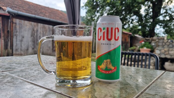 Ölglas och burk på ett marmorbord utomhus, med en träbyggnad och träd i bakgrunden.