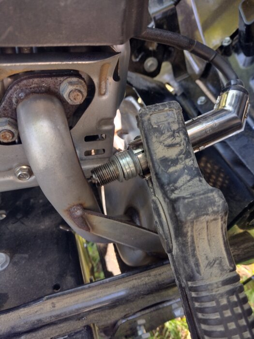 Närbild av en motor med ett tändstift och en metallsockel ansluten till en klämma. En del av motorstrukturen är synlig i bakgrunden.