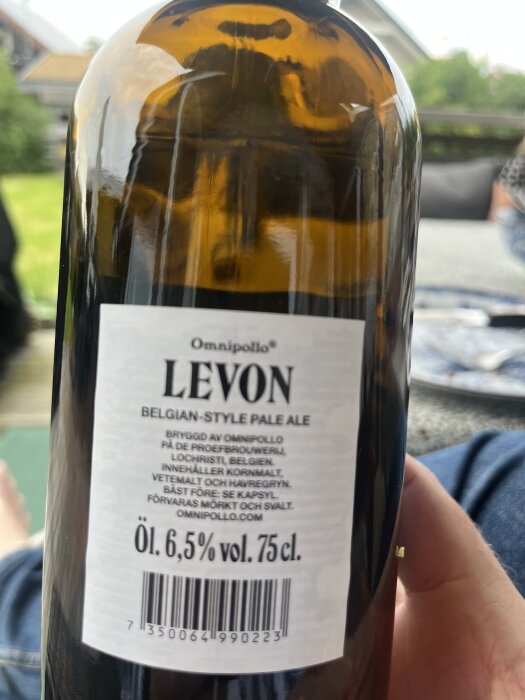 Flaska med Omnipollo Levon Belgian-style pale ale, volym 75 cl, alkoholhalt 6,5%, hålls av person på uteplats.