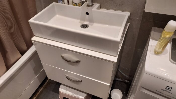 Vitt tvättställ med två lådor under där fogmassan bakom har släppt. Intill står ett badkar med brun duschdraperi och en tvättmaskin.