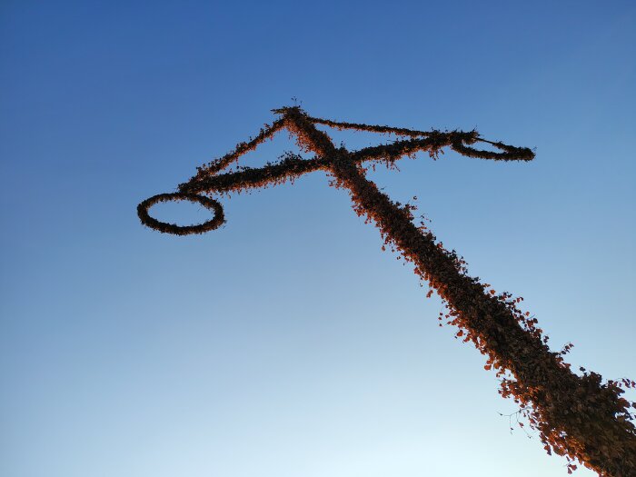 Majstång täckt med löv och blommor mot klarblå himmel.
