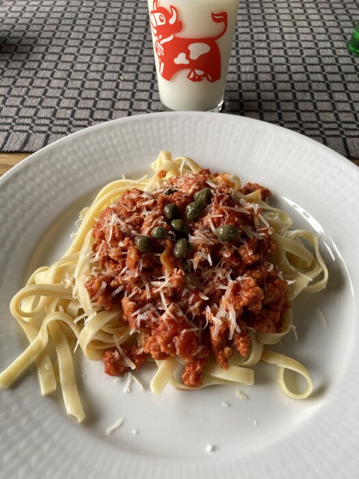 En tallrik med salsicciapasta toppad med parmesan och kapris, bredvid ett glas mjölk.