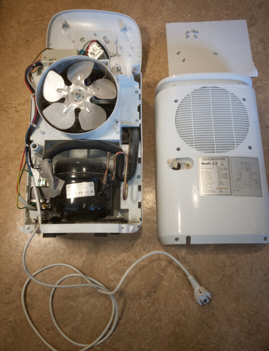 Delar av en demonterad avfuktare, inklusive fläkt och kompressor, visas på en bänkskiva. Locket och några skruvar ligger bredvid.