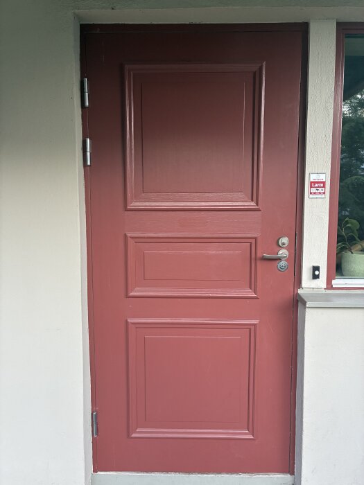 Röd trädörr med tre paneler och metallhandtag, justerad för att åtgärda hängproblem. Karmen filmas delvis; litet fönster synligt bredvid.