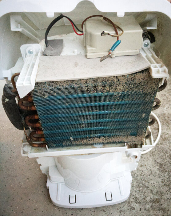 Avfuktarens invändiga komponenter efter demontering, visar kylslingor med damm och smuts samt enheten ovanpå med elektriska anslutningar.