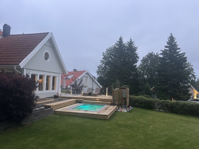 Pool med trätrall på gräsmatta utanför ett hus, omgiven av buskar och träd, med fokus på den nyinstallerade poolen.