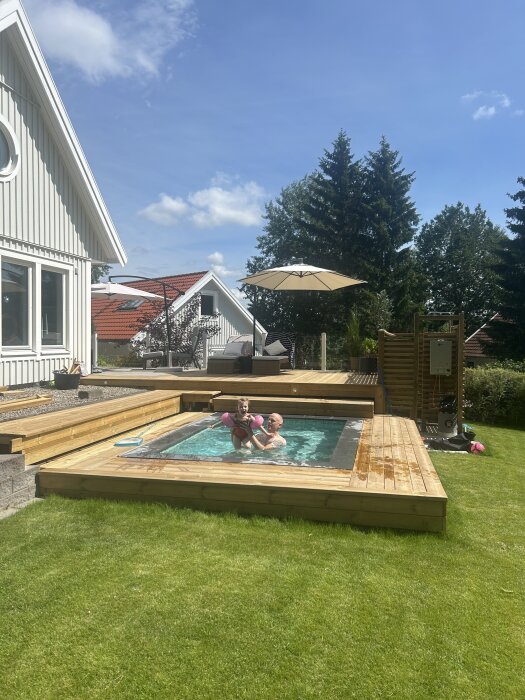 Far och dotter leker i en liten pool omgiven av trätrall i en solig trädgård. En parasoll och husets exteriör syns i bakgrunden.
