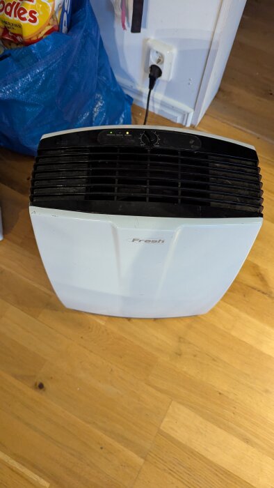 Vit Fresh luftavfuktare på trägolv, med lysande indikatorer och ansluten kontakt i väggen.