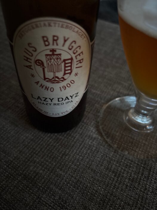 Flaska och glas med Åhus Bryggeri Lazy Dayz hazy Red IPA, fotografi på bordsyta.