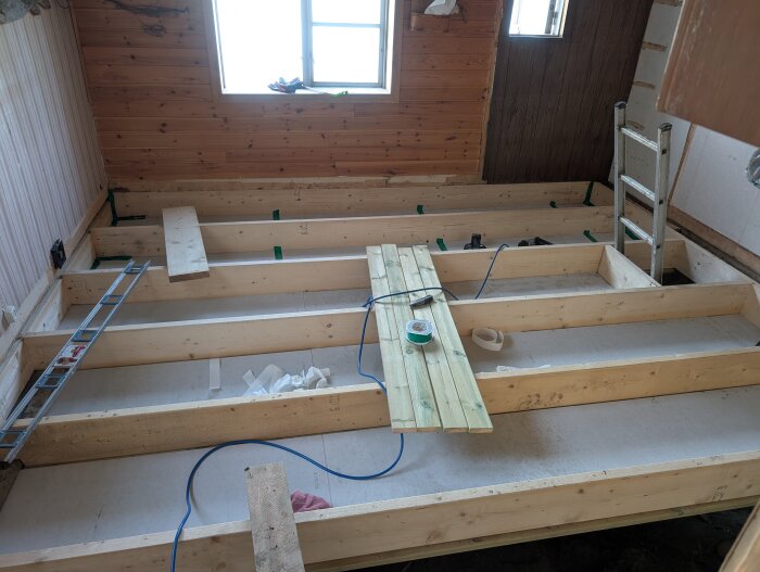 Rum under konstruktion med bjälklag och reglar; stege lutad mot väggen och träskivor med byggmaterial längs golvet.