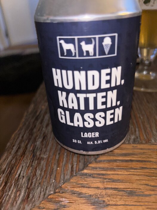 Ölburk med texten "Hunden, Katten, Glassen Lager" och symboler för en hund, katt och glass, på ett träbord.