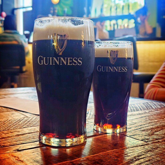 Två glas Guinness öl på träbord på en bryggeripub, med suddig bakgrund av besökare på Guinness Open Gate Brewery i Dublin.