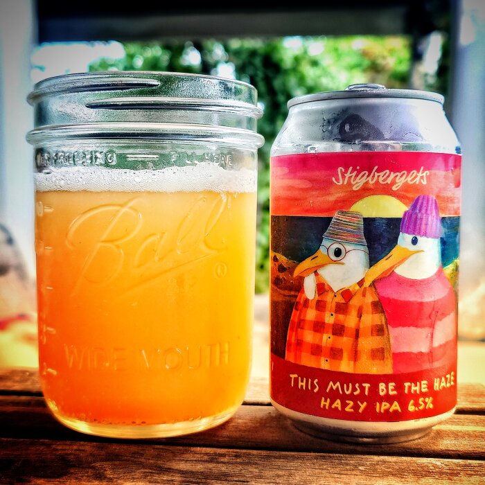 En burk Stigbergets "This Must Be the Haze" Hazy IPA 6.5% står bredvid ett glas med ljus öl på ett bord utomhus.