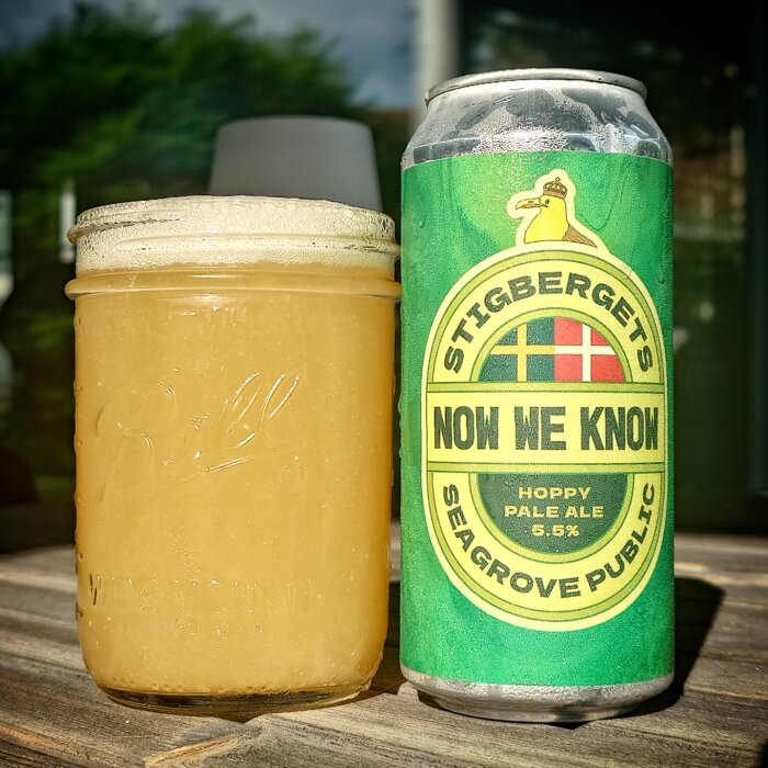Ölburk med etiketten "Stigbergets Now We Know Hoppy Pale Ale" bredvid ett fyllt glas, stående på ett träbord utomhus.