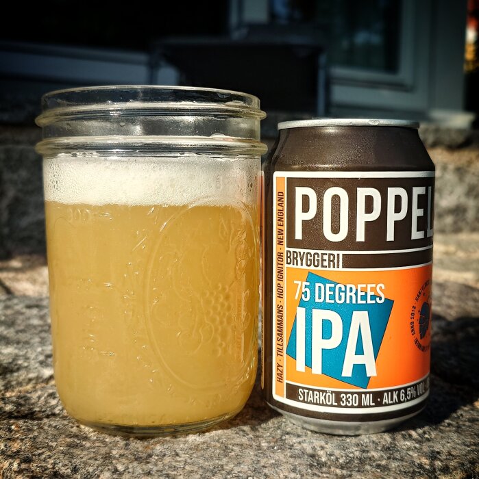 Ölglas med skummande öl bredvid en burk Poppels Bryggeri 75 Degrees IPA på stenunderlag i solljus.